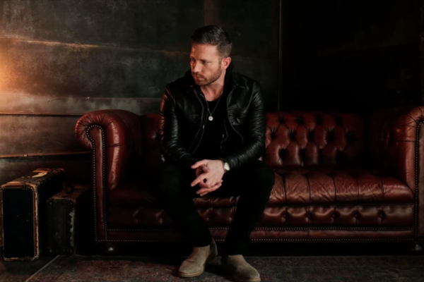 A man in a black leather jacket and boots sits on a brown tufted leather couch in a dimly lit room with dark walls. An old suitcase is on the floor nearby. He is looking to the side with his hands clasped.