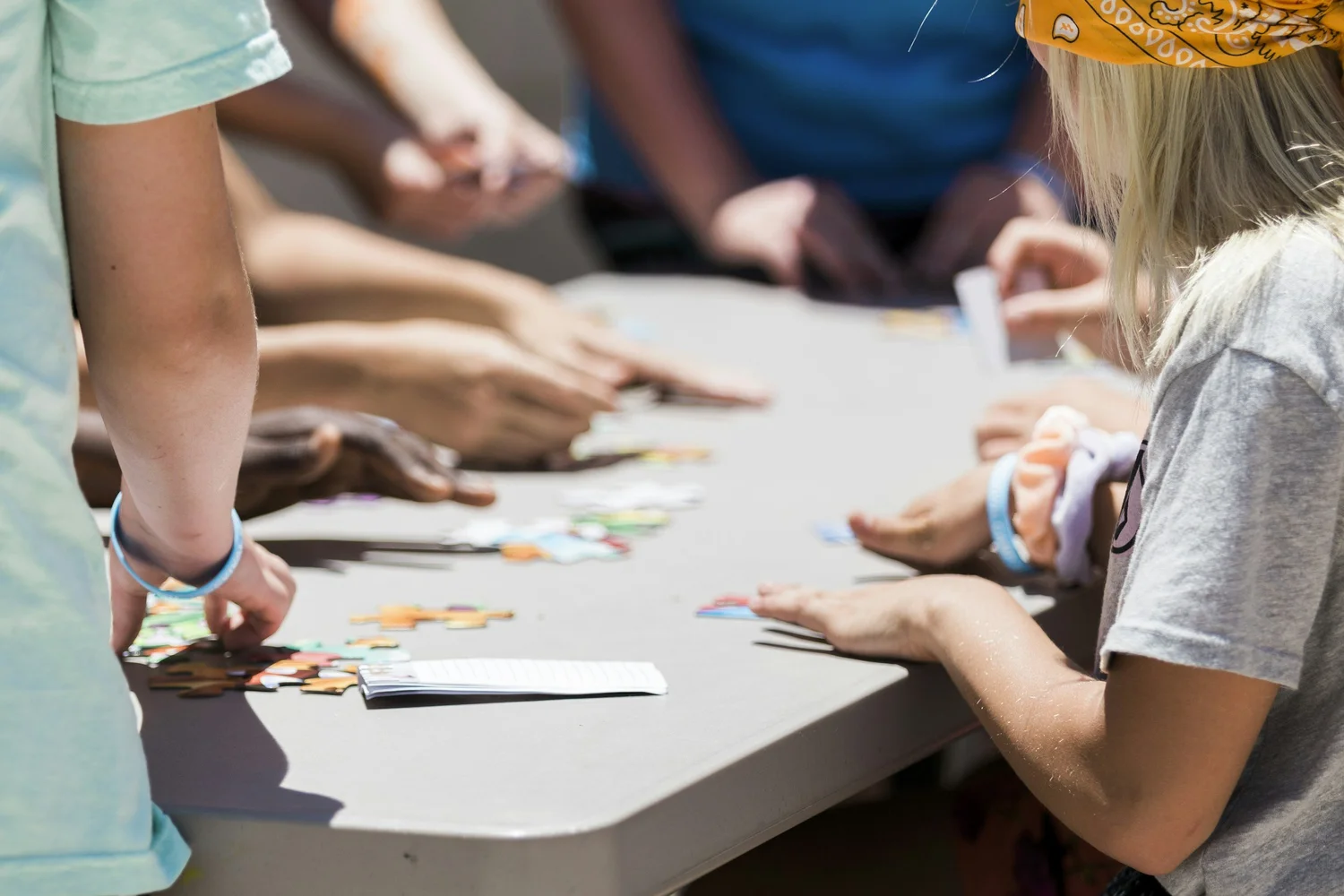 Several people gather around a table, working together to piece together a jigsaw puzzle. Only their arms and hands are visible. The surface is covered with scattered puzzle pieces and a small notepad. The setting appears to be outdoors.