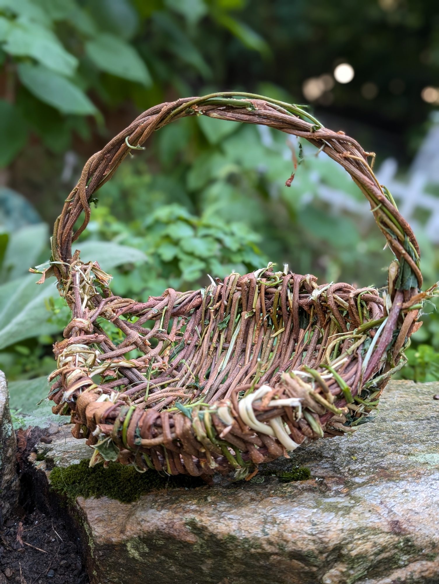 A handmade woven basket with a handle, crafted from natural twigs and vines, sits on a mossy stone outdoors. Greenery and blurred plants fill the background.