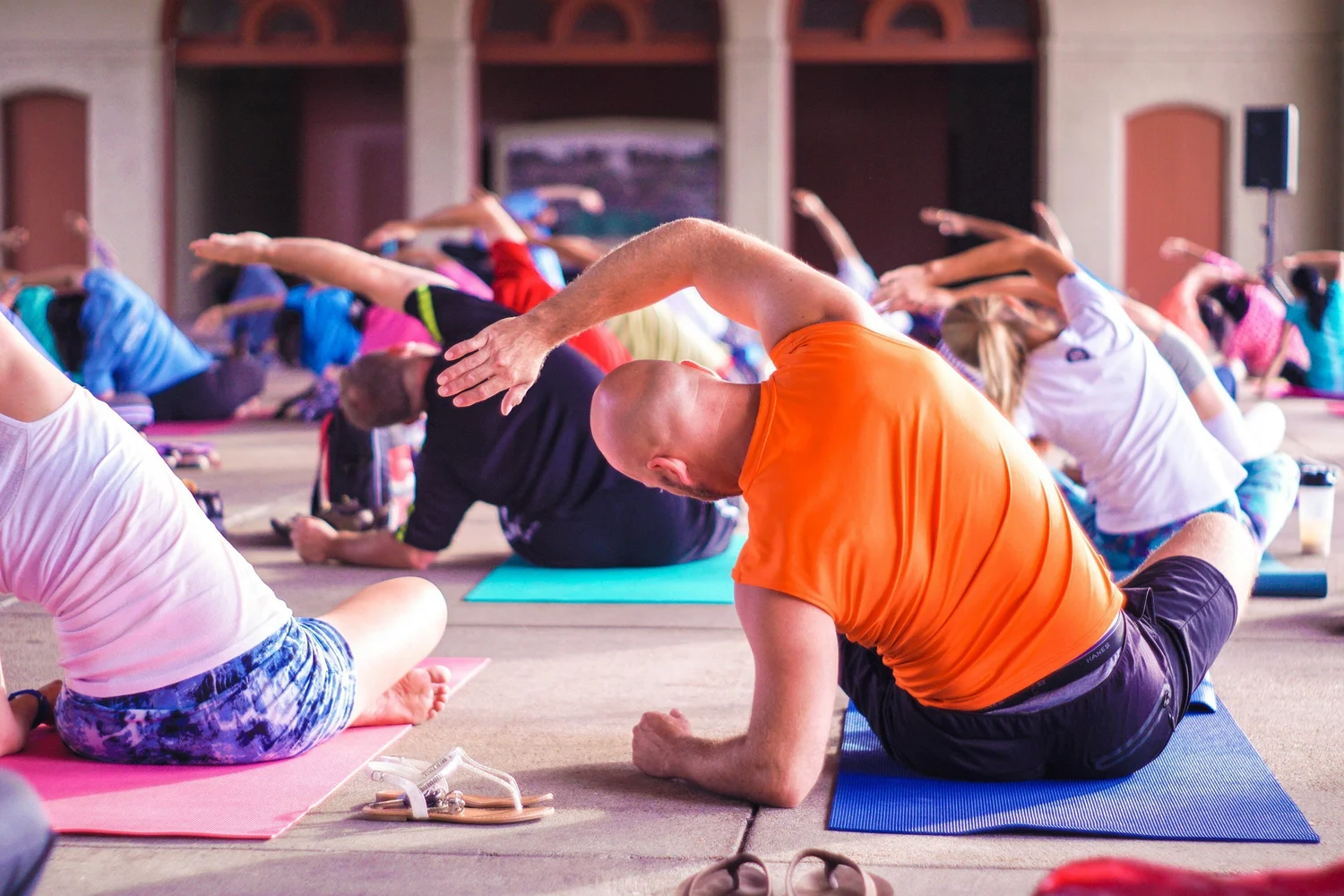A group of people are seated on yoga mats indoors, stretching and leaning to the side with one arm overhead. The setting appears to be a large hall with arched doorways in the background. The main focus is a person in an orange shirt.