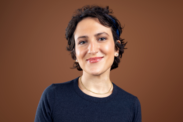 A person with short curly brown hair, wearing a dark crew-neck shirt, a gold necklace, and a blue headband, is smiling gently in front of a plain brown background.