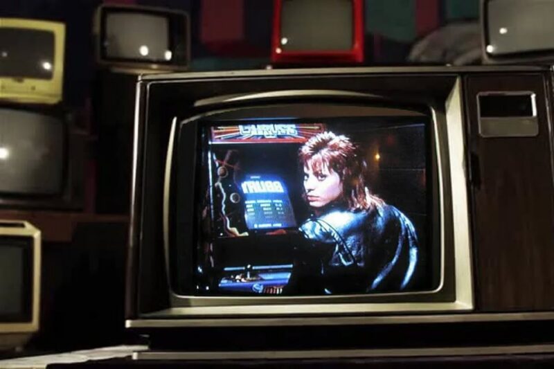 A vintage television displays an image of a person with a mullet hairstyle and a black leather jacket turning toward the camera while playing an arcade game. Several old televisions are visible in the background.