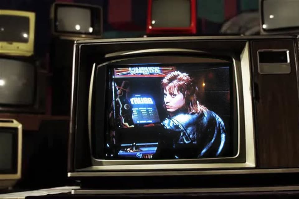 A color television displays a person with mullet hair and a leather jacket playing an arcade game, while several old televisions are stacked in the background.