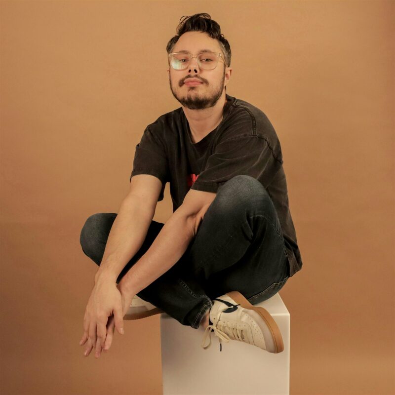 A person with short dark hair, glasses, a beard, and mustache sits cross-legged on a white cube against a plain beige background. They wear a dark T-shirt, jeans, and white sneakers, looking directly at the camera with a neutral expression.