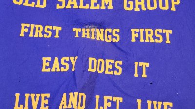 A blue fabric with yellow text reads: “OLD SALEM GROUP FIRST THINGS FIRST EASY DOES IT LIVE AND LET LIVE.” There is a small worn or damaged spot near the top center.