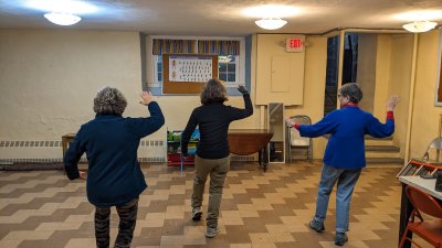 Three older adults stand in a room with tile flooring, facing away from the camera, raising their right arms as if following dance or exercise instructions. The space has beige walls, overhead lights, and a chart with letters on the far wall.