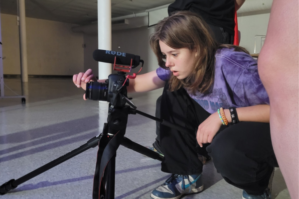 A person in a purple shirt crouches to adjust a camera with a microphone mounted on a tripod indoors. The person appears focused on the camera, with bands on their wrist, and other people are partly visible nearby.