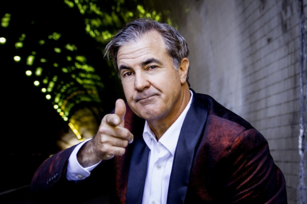 A man with gray hair, wearing a white shirt and a dark suit jacket, sits in a tunnel with brick walls and green lights in the background. He is looking at the camera and pointing, with a neutral facial expression.