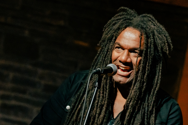 A person with long dreadlocks stands in front of a microphone, smiling while performing. The background is dimly lit with a stone or brick wall visible. The person is wearing a dark jacket.