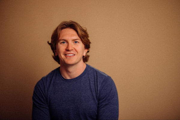 A person with shoulder-length light brown hair wearing a blue shirt sits and smiles at the camera against a plain beige background.