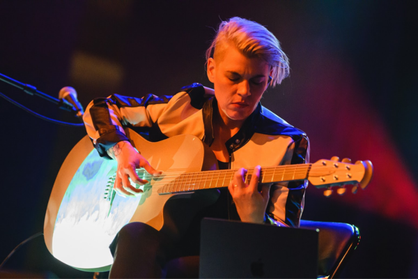 A person with short blond hair plays an acoustic guitar on stage. They are wearing a black and white jacket and sitting under colorful stage lighting. A microphone and a laptop are visible nearby.