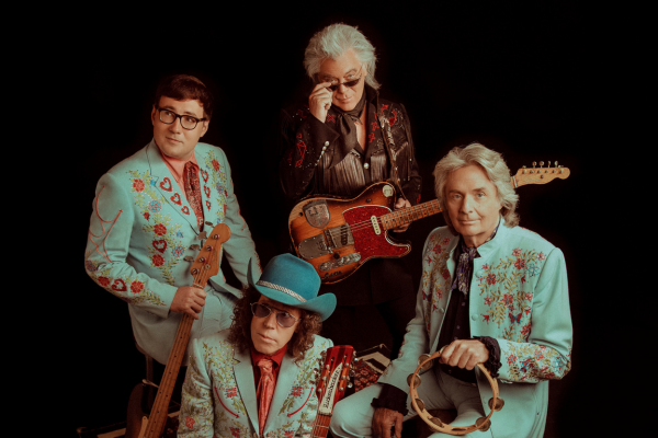 Four men in embroidered suits pose with musical instruments including guitars and a tambourine against a dark background. One man wears a blue hat, while another adjusts his glasses. Their clothing features floral and decorative patterns.