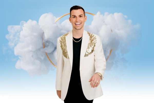 A man wearing a white blazer with gold sequin details and a black shirt stands smiling against a blue background with white clouds and a gold ring design.