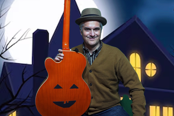 A man wearing a brown hat and sweater holds an orange guitar with a jack-o’-lantern face, sitting in front of a cartoon-style house at night with a full moon and yellow-lit windows.