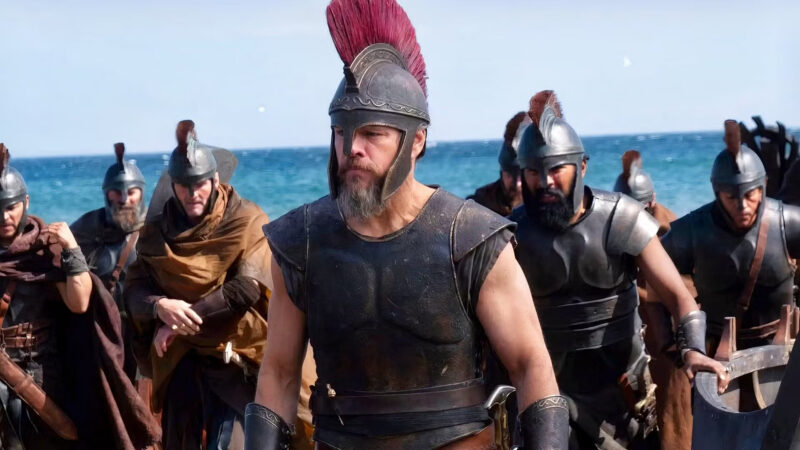A group of armored soldiers with helmets and red crests stand on a beach. The man in front, wearing leather armor and a red plume, leads the group. The sea and sky are visible in the background.