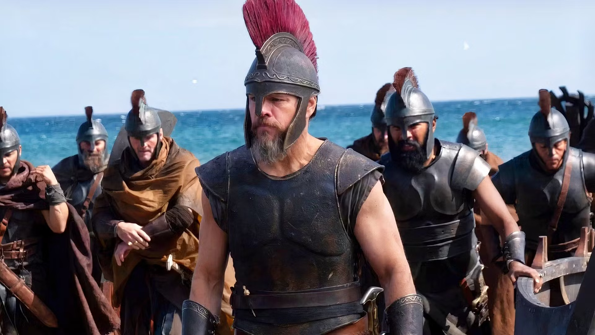 A group of armored soldiers with helmets and red crests stand on a beach. The man in front, wearing leather armor and a red plume, leads the group. The sea and sky are visible in the background.