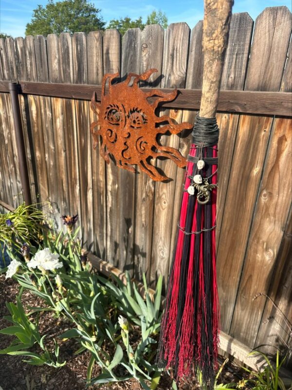 A metal sun face decor hangs on a wooden fence above blooming flowers. Next to it, a Witch City Broom with charms tied around the handle leans upright against the fence, adding magic to this charming garden scene.