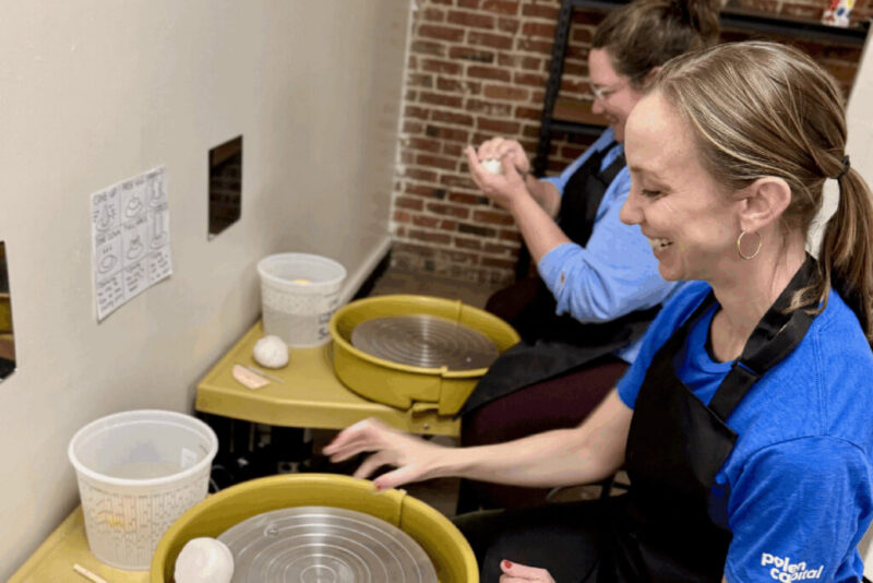 Two people sit at pottery wheels, preparing clay to throw pottery. Each has a lump of clay and wears an apron. There are plastic containers on the tables, a brick wall in the background, and a diagram is taped to the white wall beside their stations.