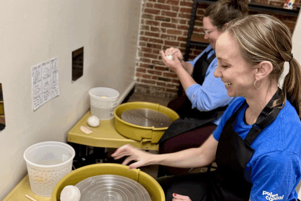 Two people sit at pottery wheels, preparing clay to throw pottery. Each has a lump of clay and wears an apron. There are plastic containers on the tables, a brick wall in the background, and a diagram is taped to the white wall beside their stations.