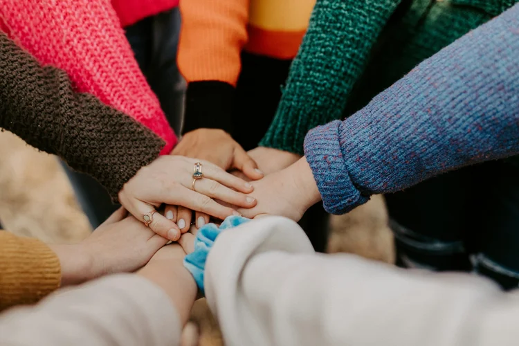 A group of people stands in a circle and places their hands on top of each other in the center. They are wearing colorful sweaters, and only their hands, arms, and parts of their torsos are visible. The background is out of focus.