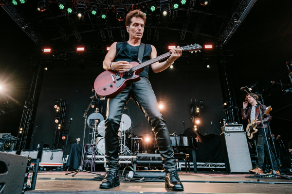 A musician wearing black clothing plays an electric guitar on an outdoor stage. Bright stage lights and equipment are visible in the background, and another band member stands to the right, also playing an instrument.