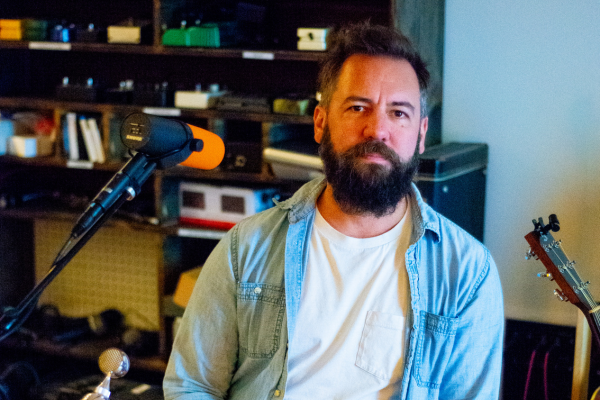 A bearded man in a white t-shirt and denim shirt sits beside a microphone in a room with shelves holding audio equipment, books, and a guitar. The lighting is warm, and the man looks directly at the camera with a neutral expression.