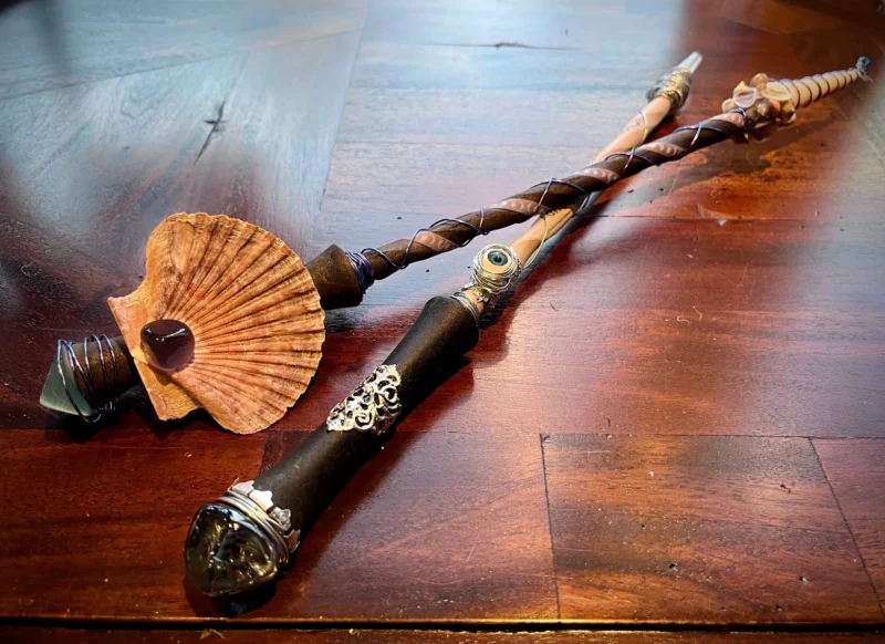Two handcrafted magic wands, perfect for a Craft Witch or Witch Session, rest on wood. One features a shell, crystal, and leather; the other boasts a black handle with metal accents and a clear crystal tip. Both are ornate and detailed.