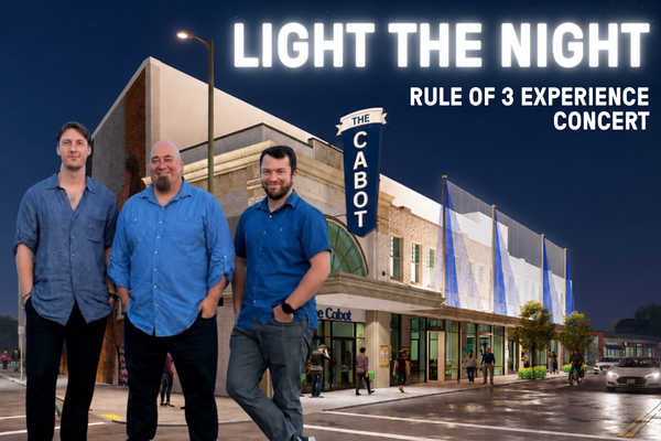 Three men stand in front of The Cabot theater at night. The marquee reads “LIGHT THE NIGHT RULE OF 3 EXPERIENCE CONCERT.” The theater is illuminated, and people are visible entering and walking nearby.