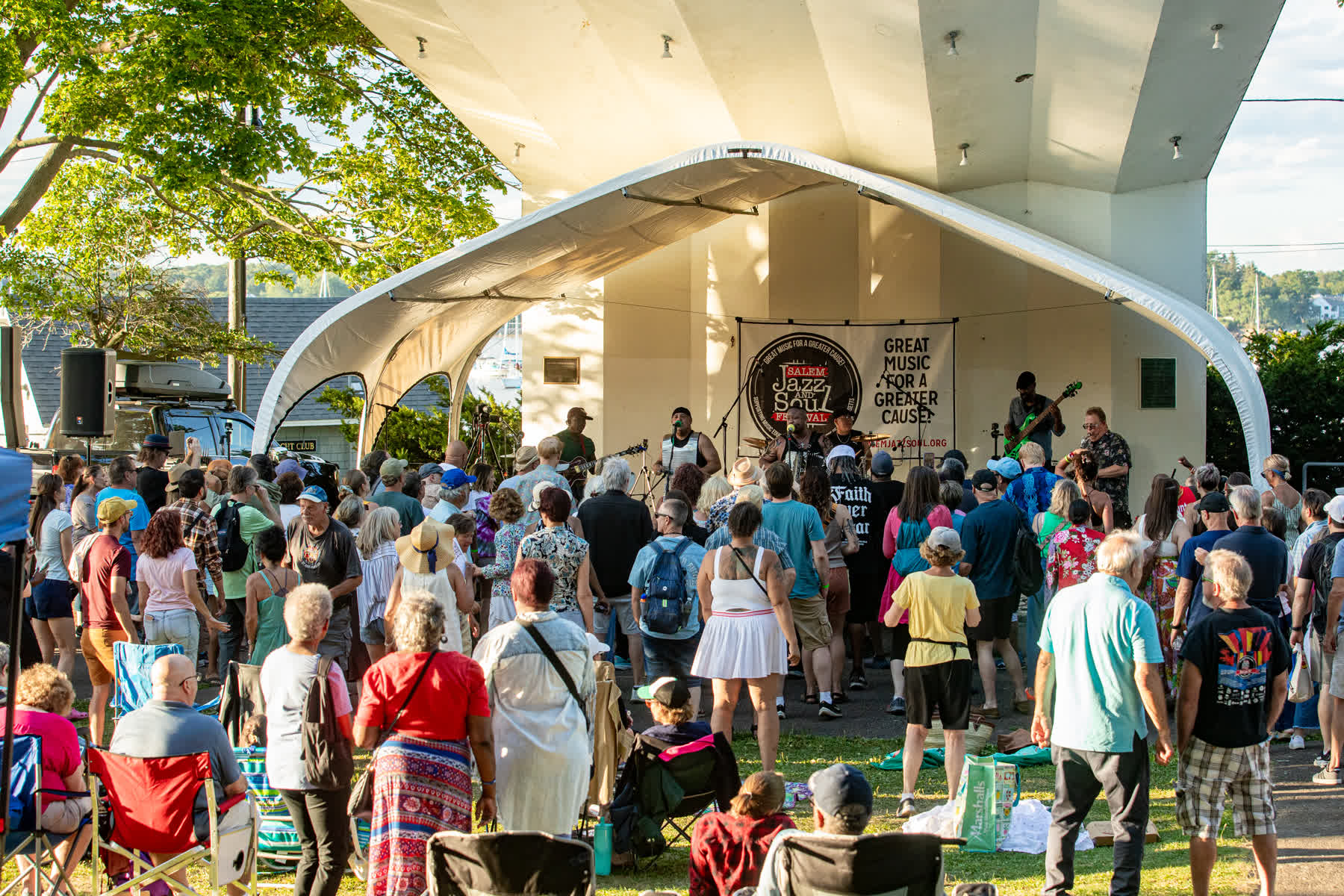 A large crowd gathers outdoors at Salem Willows in front of a white stage where a band performs for the 2026 Salem Jazz and Soul Festival. People are standing, sitting in chairs, and enjoying the music under a clear, sunny sky.