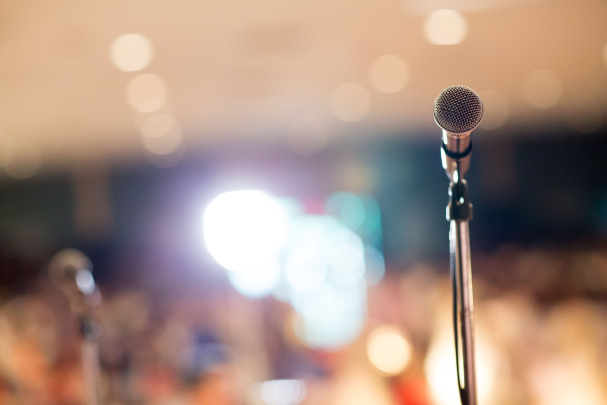 A close-up of a microphone on a stand with a blurred audience and bright lights in the background, suggesting a stage or public speaking event.
