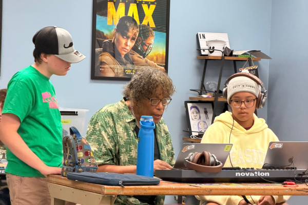 Three people are working at a table with laptops and a keyboard. One stands while two sit, focusing on their screens. A “Mad Max” poster and shelves with books and headphones are in the background. A blue water bottle is on the table.