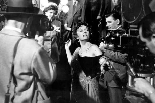 A woman in an elaborate dress stands in the center as cameras and crew surround her on a film set. The scene is in black and white, capturing a dramatic moment with several people focused on the filming process.