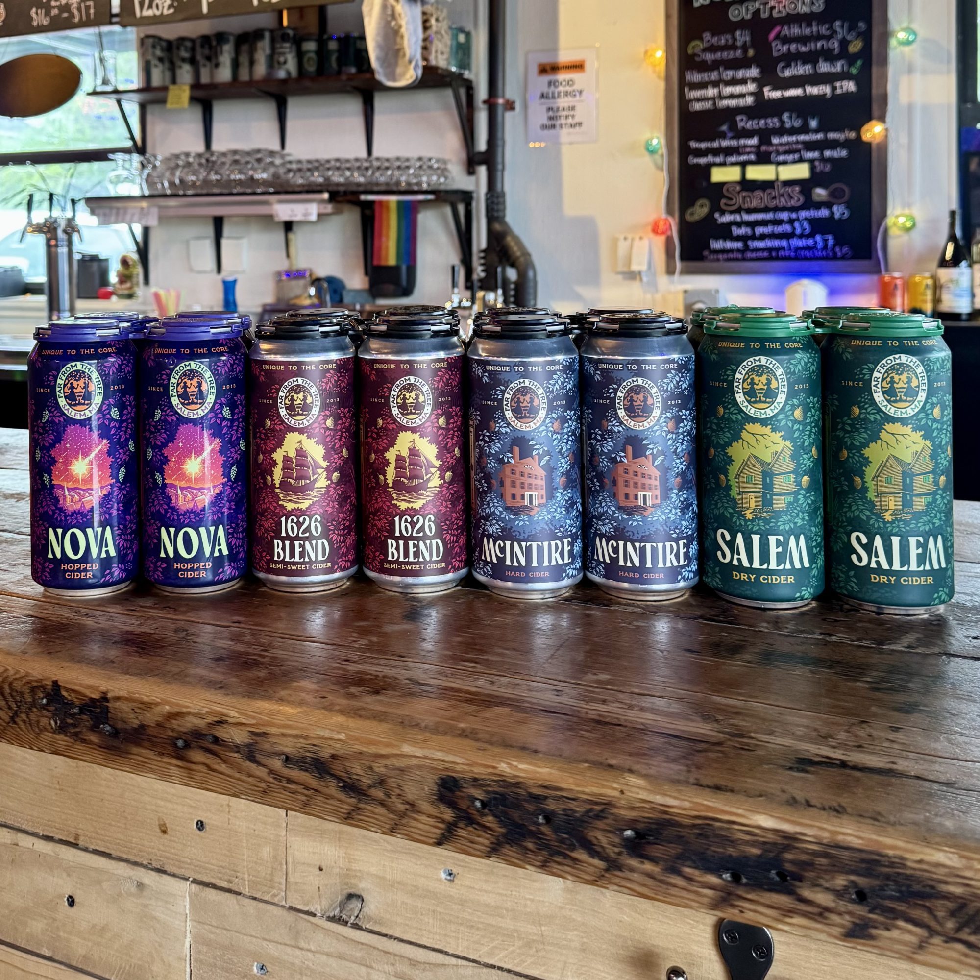 Eight cans of hard cider are lined up on a wooden counter in a bar. The cans are labeled Nova, 1626 Blend, MacIntyre, and Salem, with two cans of each flavor. A bar area and a chalkboard menu appear in the background.