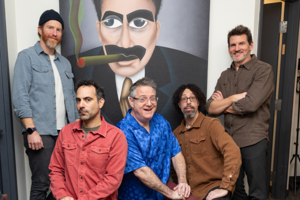Five men pose indoors in front of a large painting of a man with glasses and a cigar. Three men are seated in front, while two stand behind them. Everyone is casually dressed. The setting appears to be an office or studio.