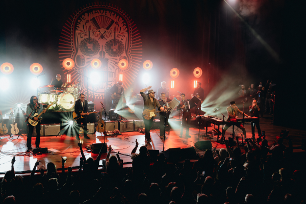 A band performs onstage with guitars, drums, keyboards, and microphones. Bright stage lights and a large backdrop with circular designs are visible behind them. The audience is in the foreground, some with raised arms, watching the show.