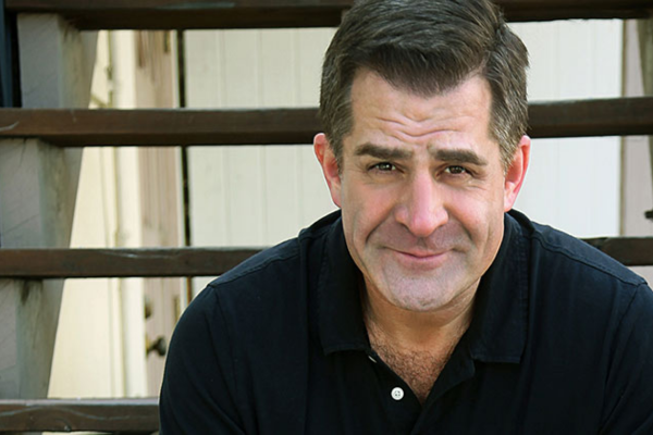 A man with short brown hair wearing a black collared shirt sits in front of wooden stairs and looks at the camera, smiling slightly. The background is out of focus.