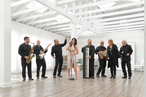 A band of seven musicians dressed in black stands in a bright, spacious room. They play guitar, saxophone, keyboard, and bass while a woman in a sparkly dress sings into a microphone at the center. Everyone appears engaged and smiling.