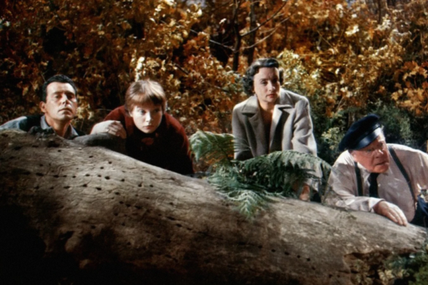 Four people—three adults and one child—crouch behind a fallen tree trunk in a forested area, looking intently ahead. The background is filled with autumn-colored foliage.