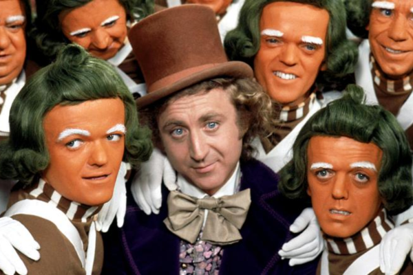 A person in a brown top hat and bow tie is surrounded by people with orange faces, green hair, and white-gloved hands, all wearing matching brown and white costumes.
