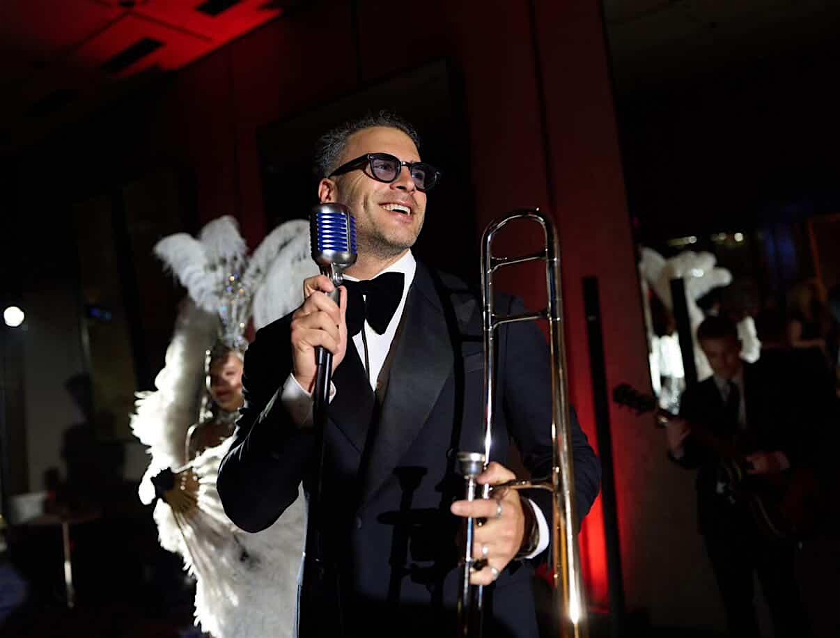 A man in a tuxedo holds a trombone and sings into a vintage microphone. A feathered performer stands in the background, and another musician with a guitar is visible in dim lighting.