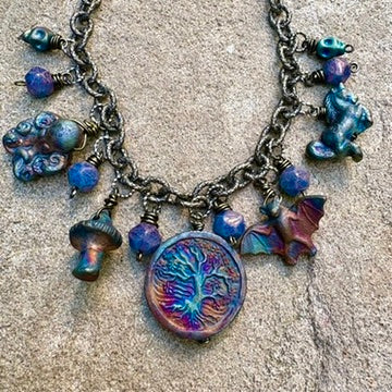 A Charm Bar necklace with metallic blue and purple charms—tree medallion, mushroom, bat, octopus, animal figure, and round beads—evokes Dark Arts style on a textured stone surface.