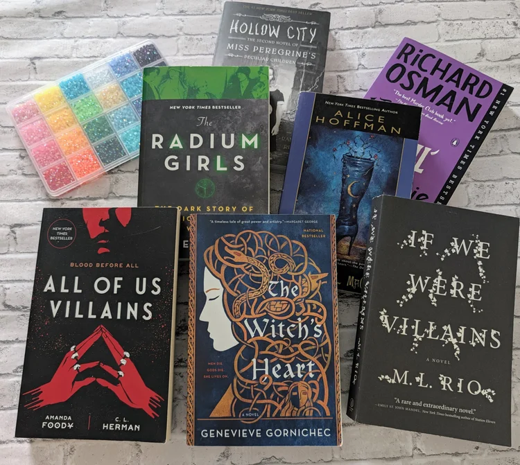 Nine books are arranged on a white surface, including titles like “The Radium Girls,” “If We Were Villains,” and “All of Us Villains,” with a colorful bead organizer placed in the top left corner.