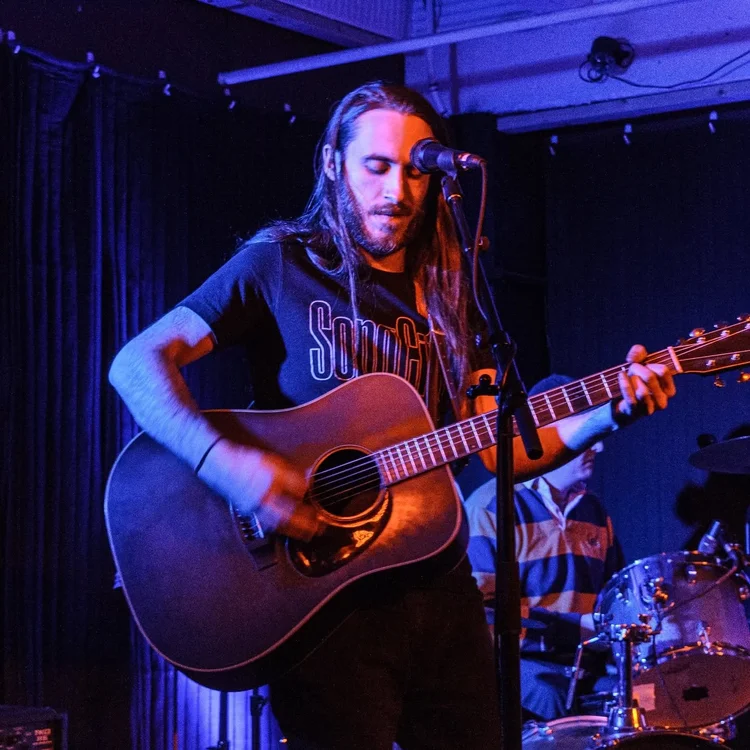 Nate Goyette with long hair plays an acoustic guitar and sings into a microphone on stage. Purple and blue lights set the mood for this live music performance, with a drum set and another musician visible in the background. No cover required.