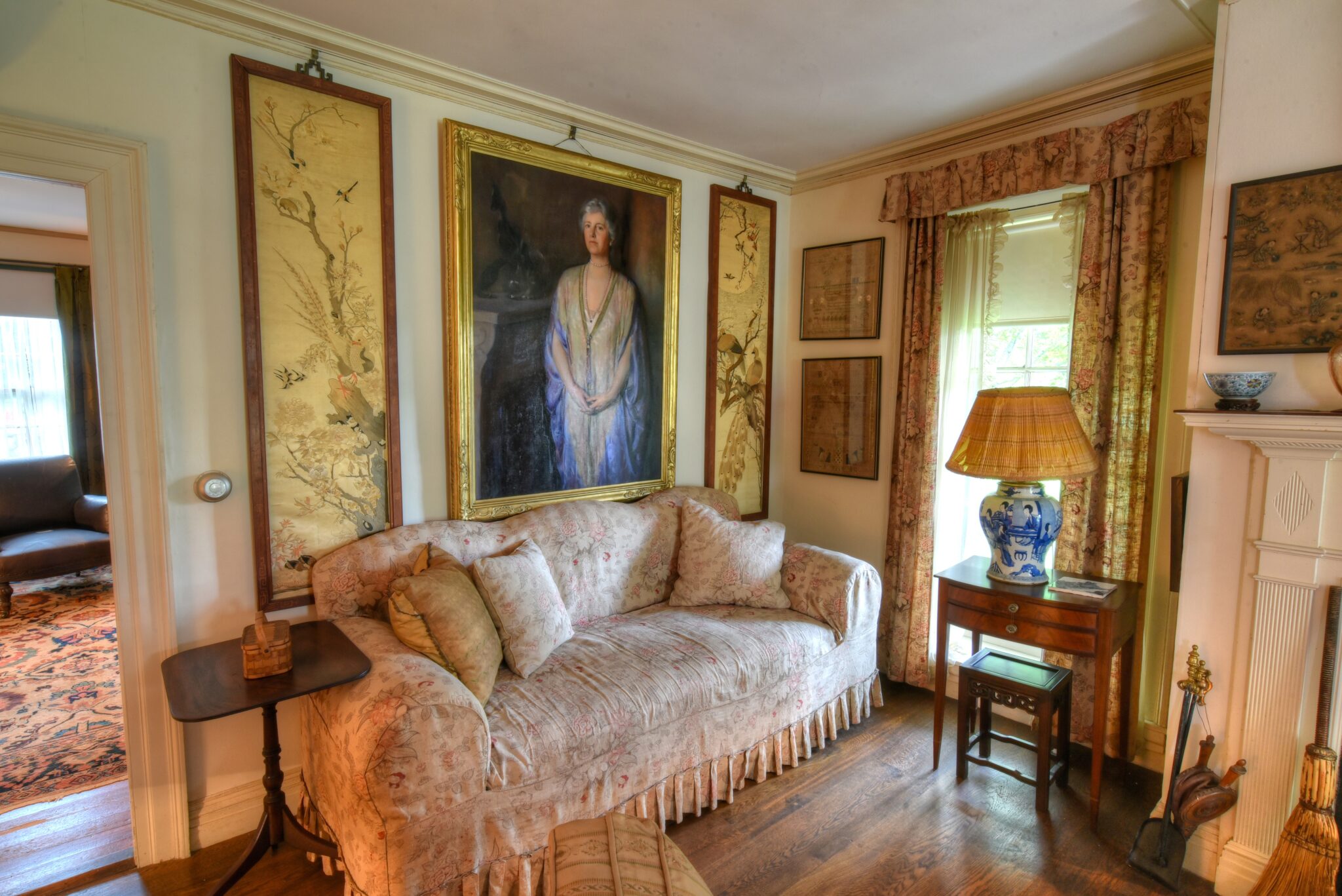 A floral-patterned sofa sits below a large portrait of a woman. Enjoy framed art and patterned curtains as if on a Stevens-Coolidge guided house tour, with hardwood flooring and vintage details offering a true passport to the past.