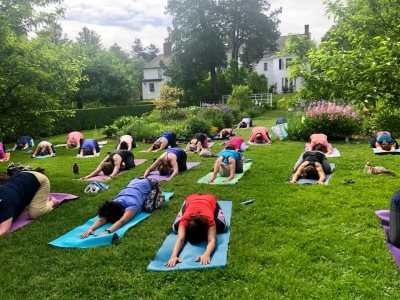 A group practices morning yoga outdoors on a grassy lawn, each on a mat, stretching with arms extended forward. Gardens, trees, and a white house are visible in the background under a partly cloudy sky.