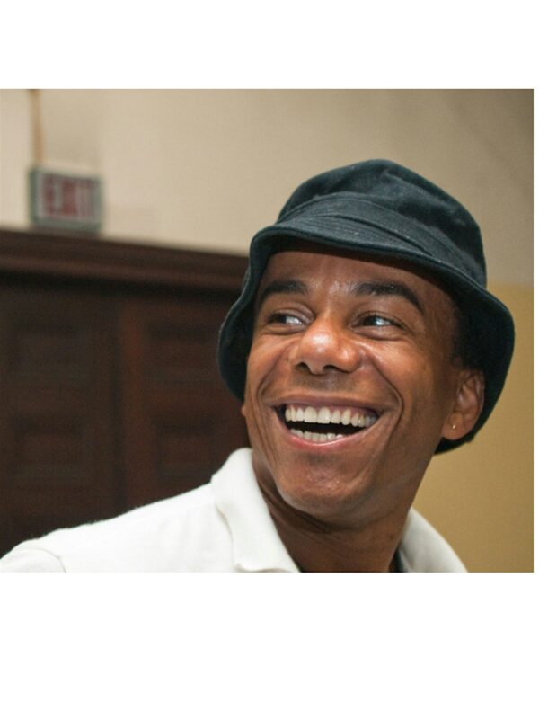 A person wearing a black bucket hat and white shirt is smiling and looking to the side. A dark wooden door and an exit sign are visible in the background.