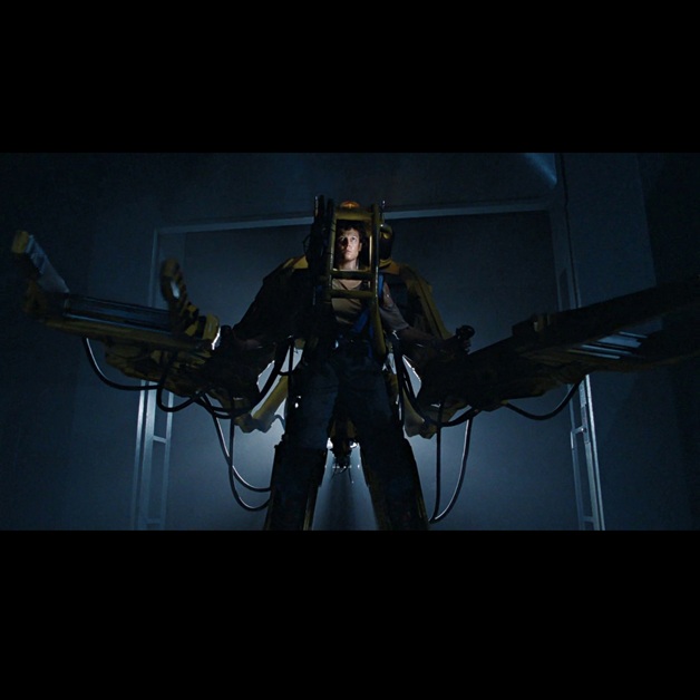 A person stands inside a large yellow mechanical exosuit with extended arms in a dimly lit room, framed by a doorway and illuminated from behind—a scene iconic to Aliens, now celebrated in the 40th Anniversary Extended Edition.