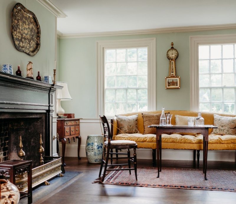 Experience this traditional living room on Self-Guided House Tours, featuring a cozy fireplace, wooden furniture, yellow upholstered bench, blue and white ceramics, natural light, and elegant rugs for a warm and inviting atmosphere.