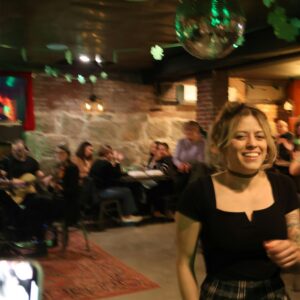 A woman with shoulder-length hair and a black top smiles in a dimly lit Essex County bar. Several people sit at tables, while green decorations and a disco ball hang from the ceiling, capturing the lively vibe of CCE Nightlife and Sevenings.