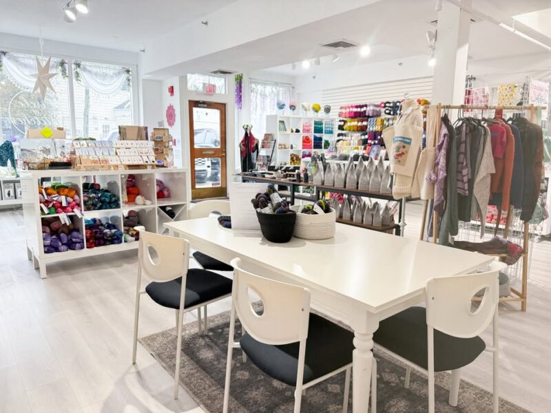 A brightly lit yarn and craft store, home of Circle of Stitches, features shelves of colorful yarn and supplies. Centered is a white table with black chairs, baskets of yarn on top, perfect for Stitch Night gatherings in the natural light by the entrance.